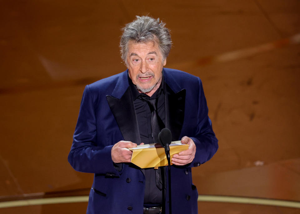 Al Pacino on stage holding an envelope at an award ceremony, wearing a dark suit with a black shirt and tie