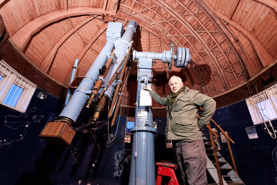 an astronomer in a green jacket and brown pants stands to the right of a giant telescope