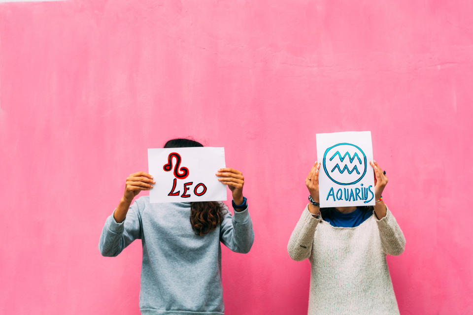 Two individuals standing against a plain background holding signs with zodiac symbols and names: Leo and Aquarius, obscuring their faces