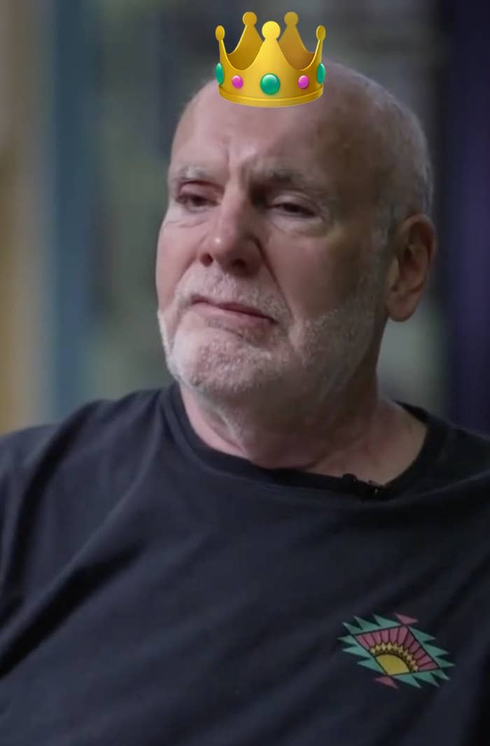 Don Vultaggio with a white beard and mustache, wearing a dark shirt with a colorful design on the chest, looks pensively off camera
