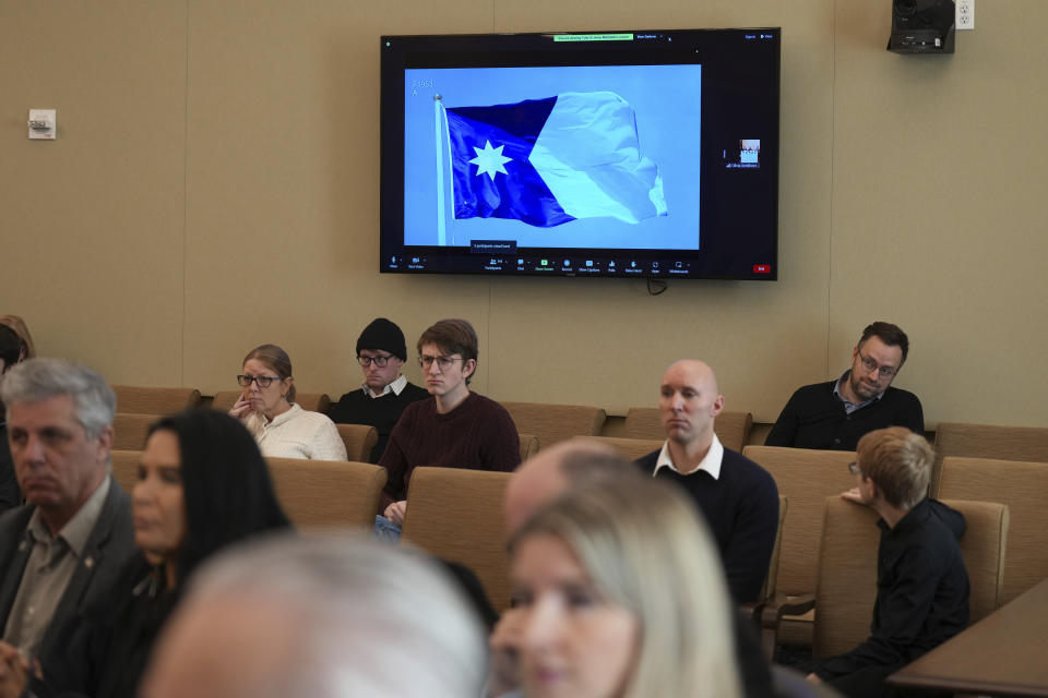 People sit in the hearing room as Minnesota's flag commission picks a final flag design, seen on the screen uesday, Dec. 19, 2023 St. Paul, Minn. The new state flag should feature an eight-pointed North Star against a dark blue background shaped like the state, with a solid light blue field at the right, a special commission decided Monday as it picked a replacement for an older design that many Native Americans considered offensive. (Glen Stubbe /Star Tribune via AP)