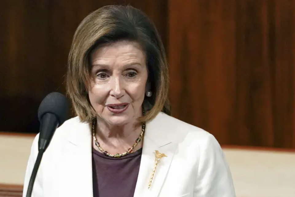 Nancy Pelosi wears a white blazer and stands in front of a microphone