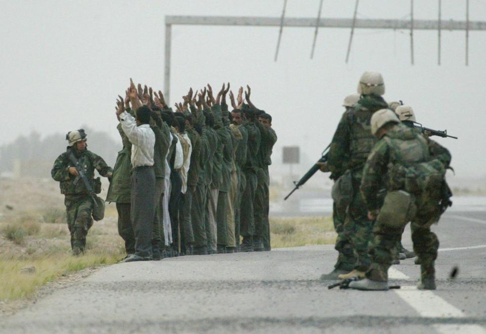 About five U.S. soldiers gather around a row of more than a dozen Iraqi soldiers standing with their hands in the air.
