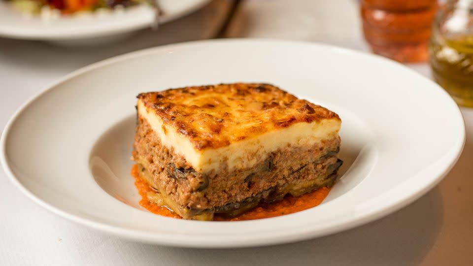 Slice of Greek moussaka served on a plate with Greek salad as a side dish white tablecloth with olive oil and vinegar in a restaurant - Jelena Jeremic/iStockphoto/Getty Images/iStockphoto