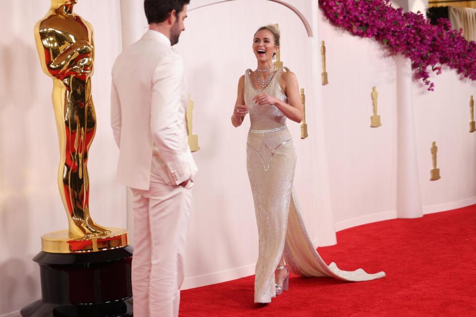 John Krasinski and Emily Blunt, both in white, laugh on the red carpet.