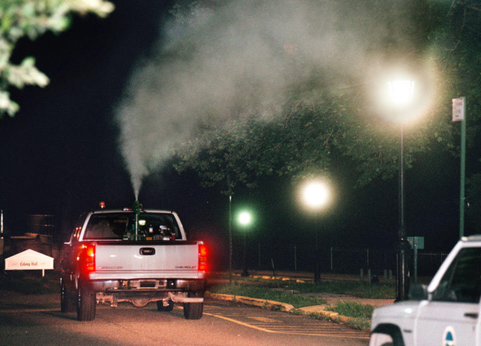 A truck emits thick smoke into the night, possibly engaging in street maintenance. Only vehicle lights and streetlamps illuminate the scene. No people visible