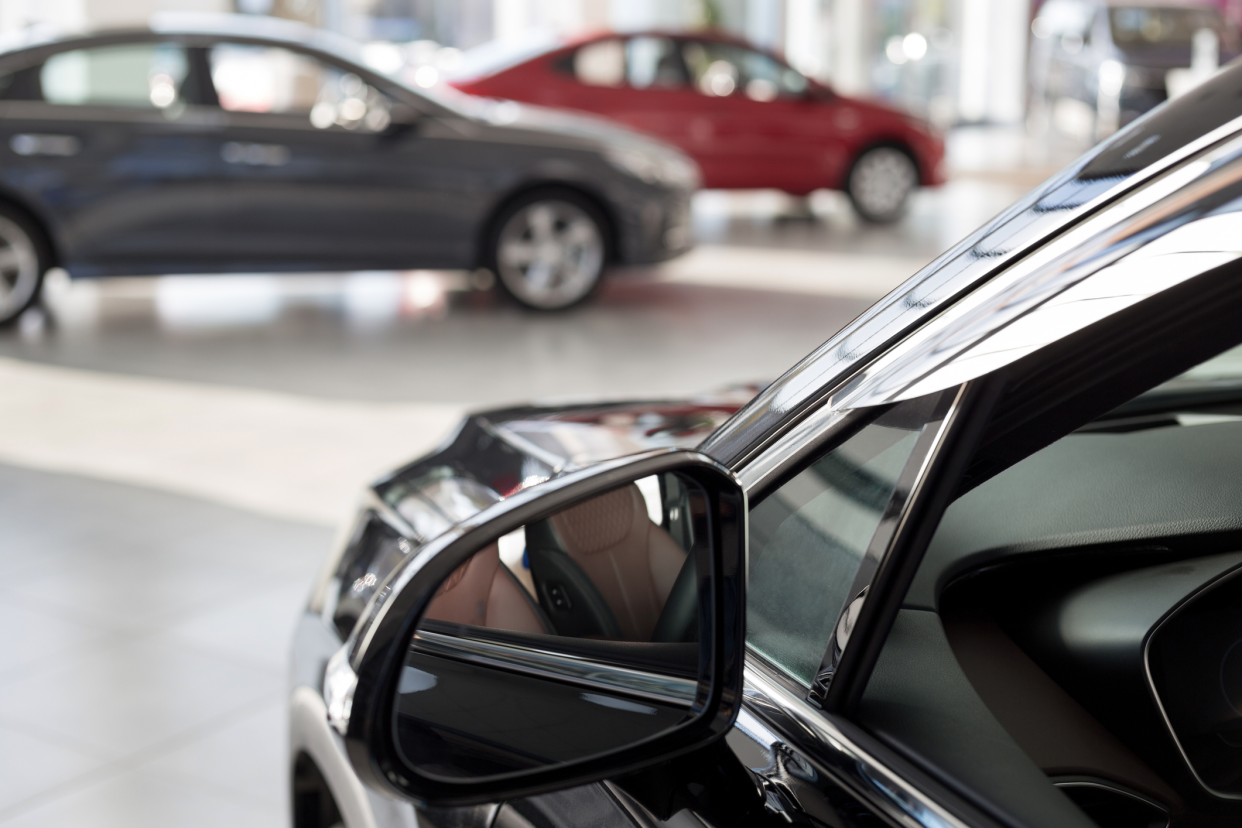 Closeup of a rear view mirror and external driver's side of a black car in a showroom of a dealership with a blurred background of two other cars in dealership