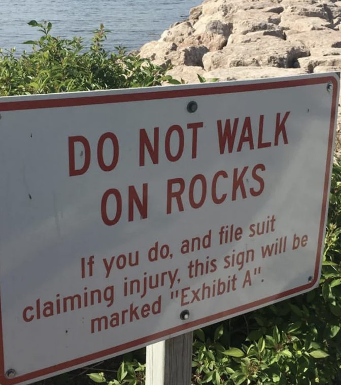 Sign near rocks reads, "DO NOT WALK ON ROCKS. If you do, and file suit claiming injury, this sign will be marked 'Exhibit A'."