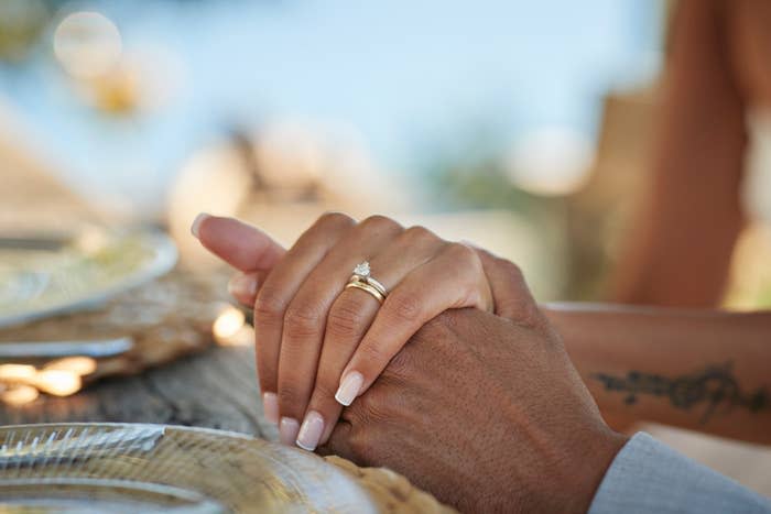 Two hands holding each other on a table, one with a wedding ring and the other with a tattoo on the wrist. Plates are visible in the foreground