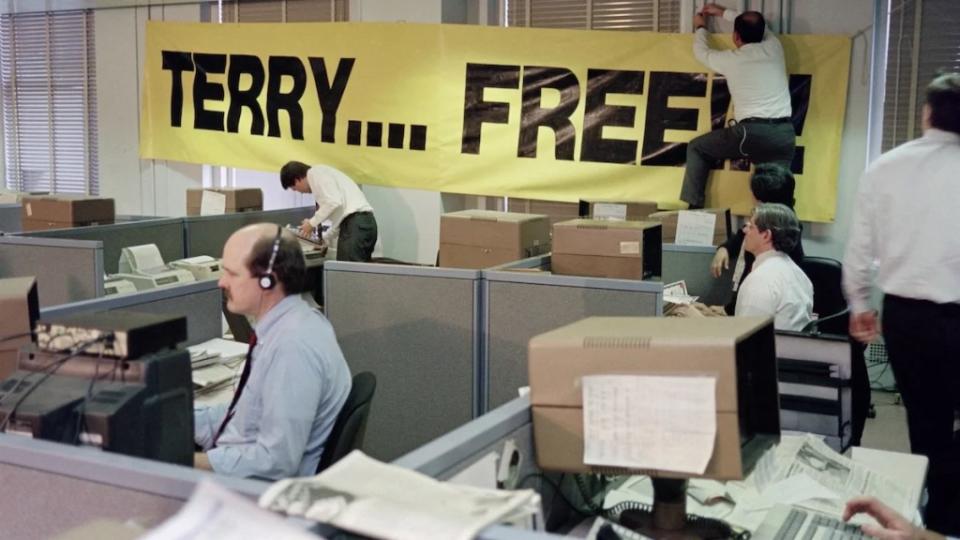A newsroom with a large banner reading "Terry... Free!" One person hangs the sign while others work around the room.
