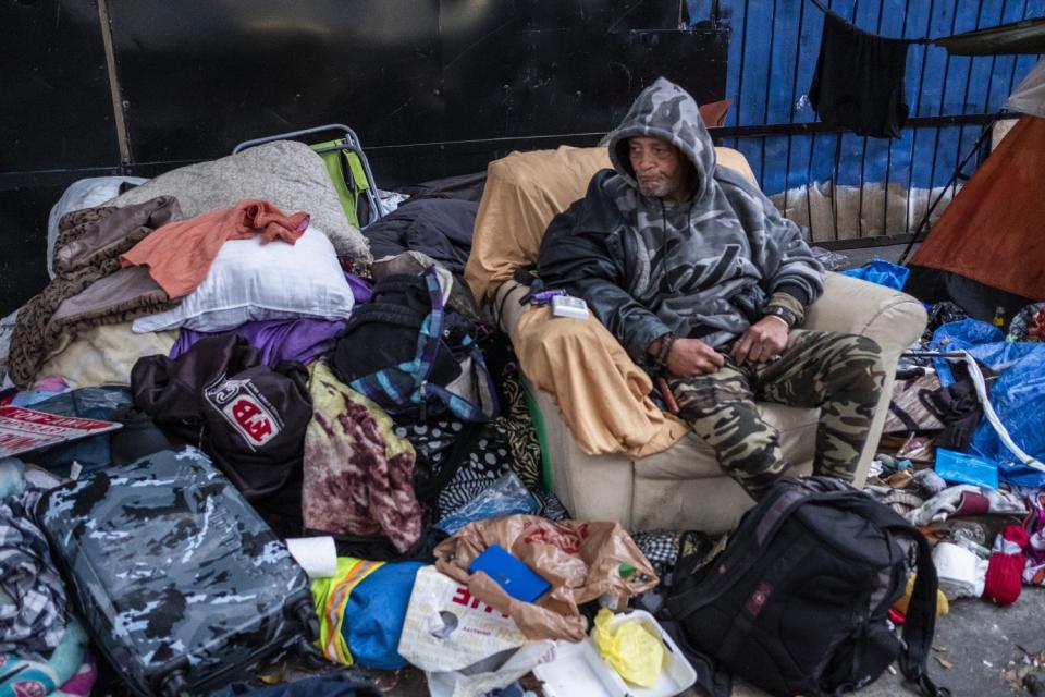 A man who goes by the name Big O sits in a chair next to mounds of clothes on the sidewalk in skid row