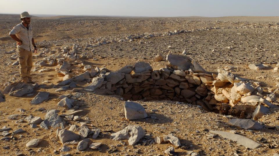 A man in a hat stands near a rock-wall pile in the desert.