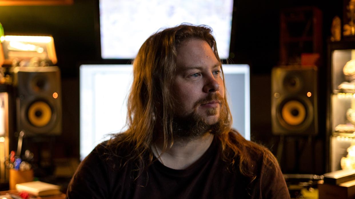  A man with long brown hair is surrounded by music equipment and two large speakers behind him. . 