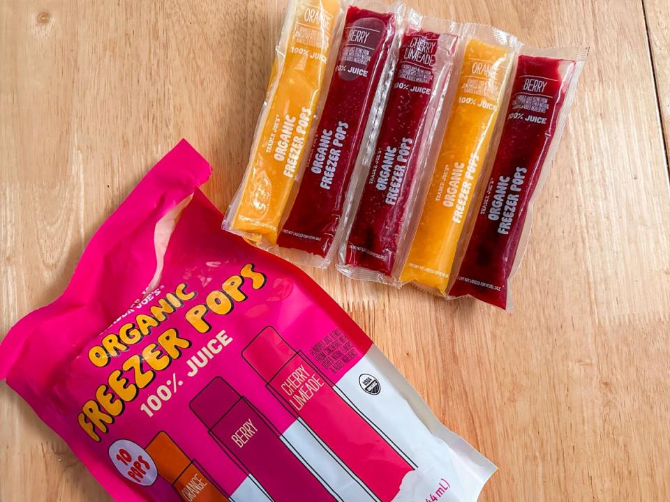 A pink and white bag with illustrations of pink, red, and orange ice pops on it sits next to orange and red ice pops in clear plastic packaging on a wooden table