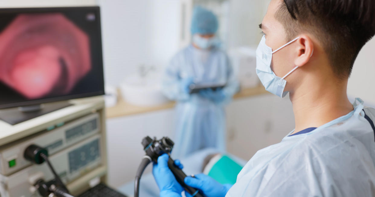  Doctor wearing a surgical mask stands in front of a monitor while performing a colonoscopy on a patient that's out of frame. A nurse in scrubs stands in the background with a tablet. 