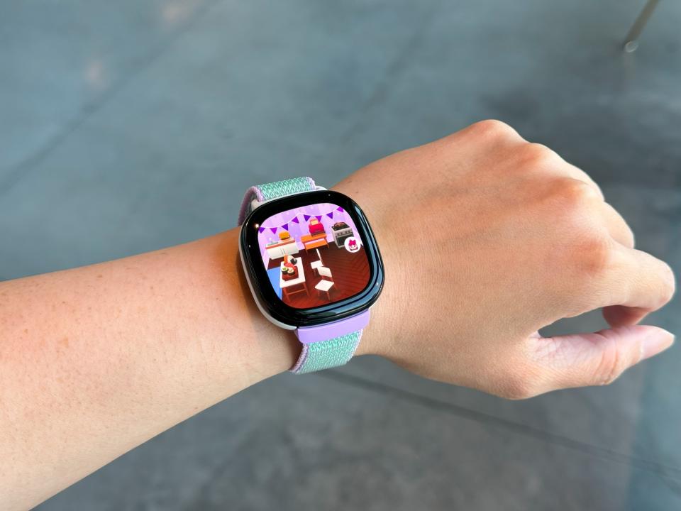 Das Fitbit Ace LTE am Handgelenk wird in die Luft gehalten, mit einem Cartoon-Raum auf dem Bildschirm.
