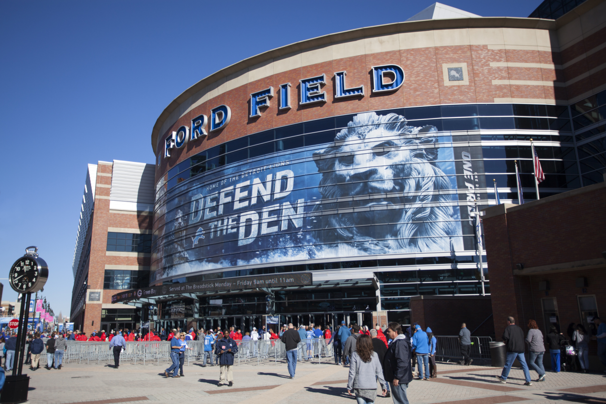 Exterior of Detroit Lions, Ford Field, Detroit, numerous people walking around and getting into lines, with a huge 'Defend the Den' blue lion sign, against a blue sky