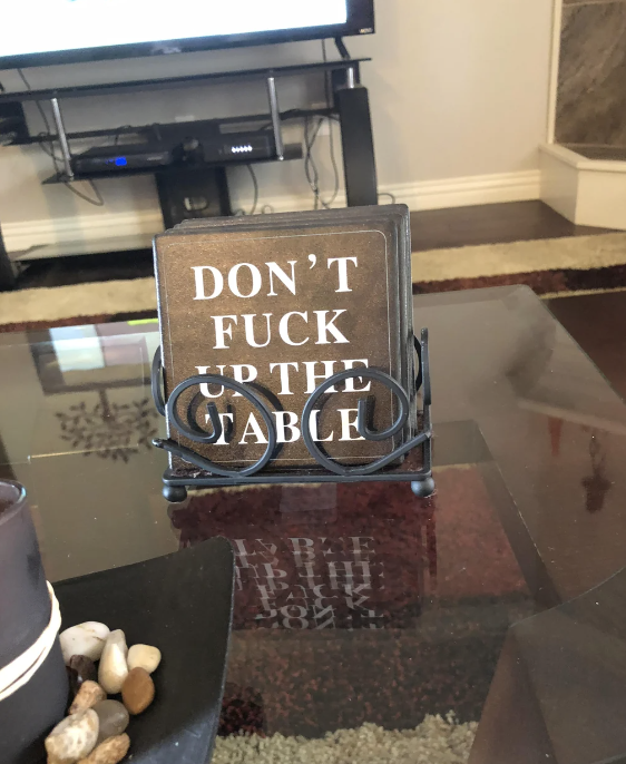 A photo of a coaster on a table that reads, "DON'T FUCK UP THE TABLE," placed in a holder. A living room setup is visible in the background