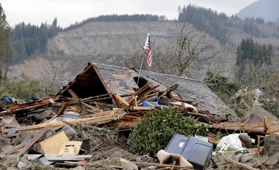 A flag sticks out of a hole in a demolished home near where a deadly mudslide from the hill behind that happened several days earlier ended, Tuesday, March 25, 2014, in Arlington, Wash. At least 14 people were killed in the 1-square-mile slide that hit in a rural area about 55 miles northeast of Seattle on Saturday. Several people also were critically injured, and homes were destroyed. (AP Photo/Elaine Thompson)