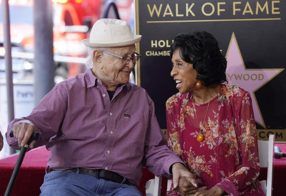 An older man and woman sit in chairs on a red carpet