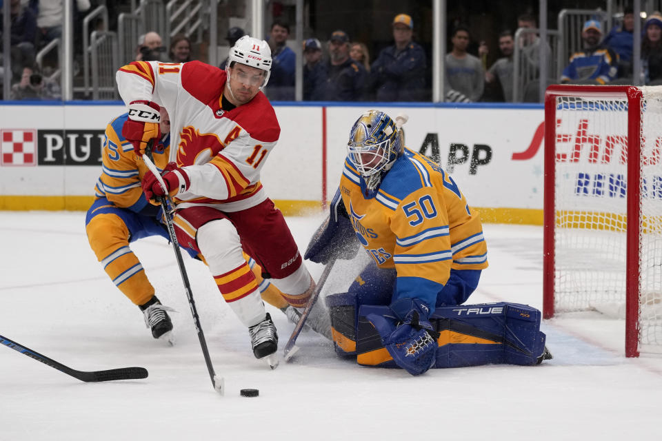 Calgary Flames' Mikael Backlund (11) reaches for a loose puck as St. Louis Blues goaltender Jordan Binnington (50) defends during the first period of an NHL hockey game Tuesday, Jan. 10, 2023, in St. Louis. (AP Photo/Jeff Roberson)