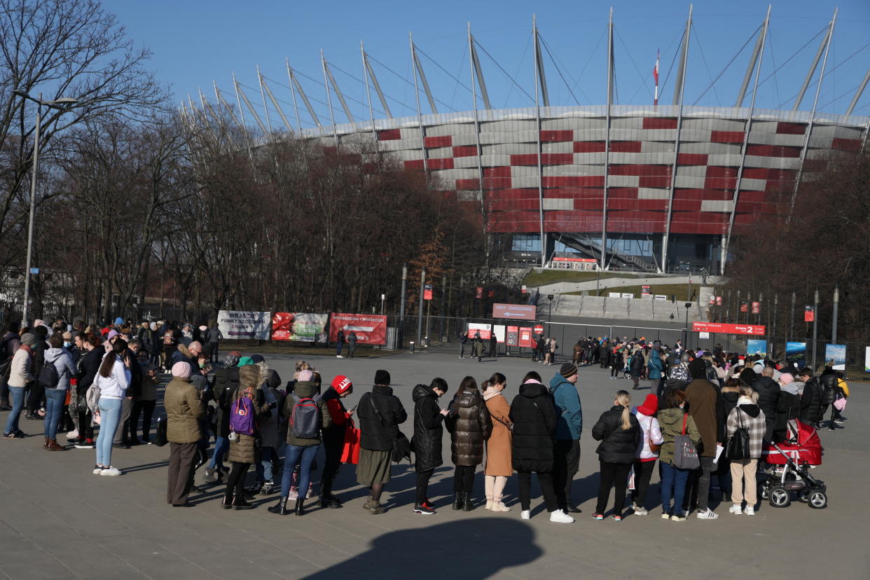 People wearing cold weather clothes stand in a long line leading to a fence next to a stadium.
