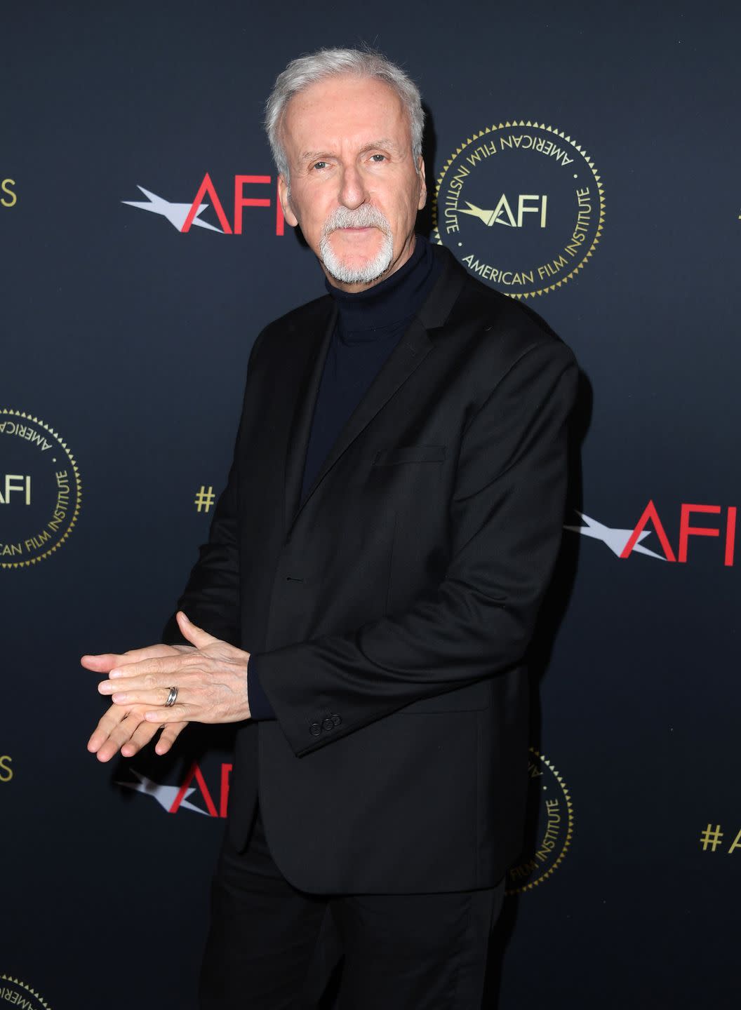 james cameron, an older man stands looking at the camera clasping his hands, he wears a black suit and turtleneck