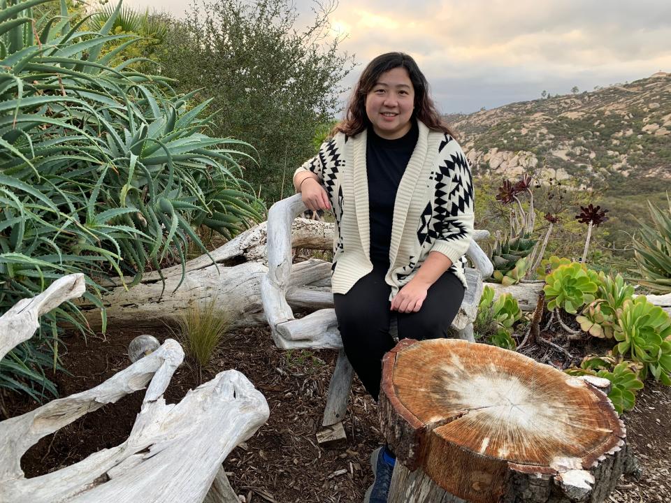 A woman sitting on a chair made of white wood in front of a hilly scenic area.