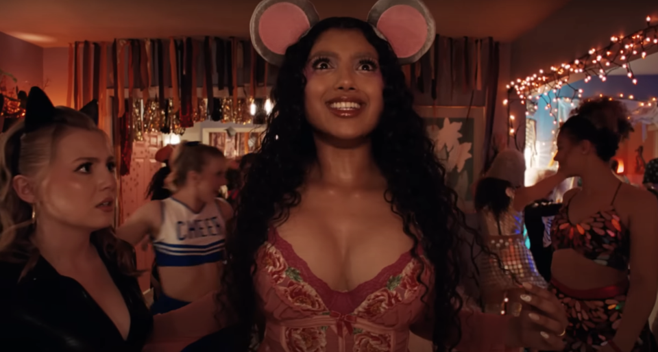 in a scene, a group of people at a lively party in costumes. Karen wears mouse ears and a floral outfit. Other guests, including a cheerleader, are seen in the background