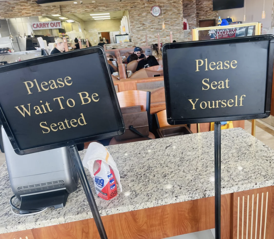 Two contradictory signs at a restaurant: one says "Please Wait To Be Seated" and the other "Please Seat Yourself."