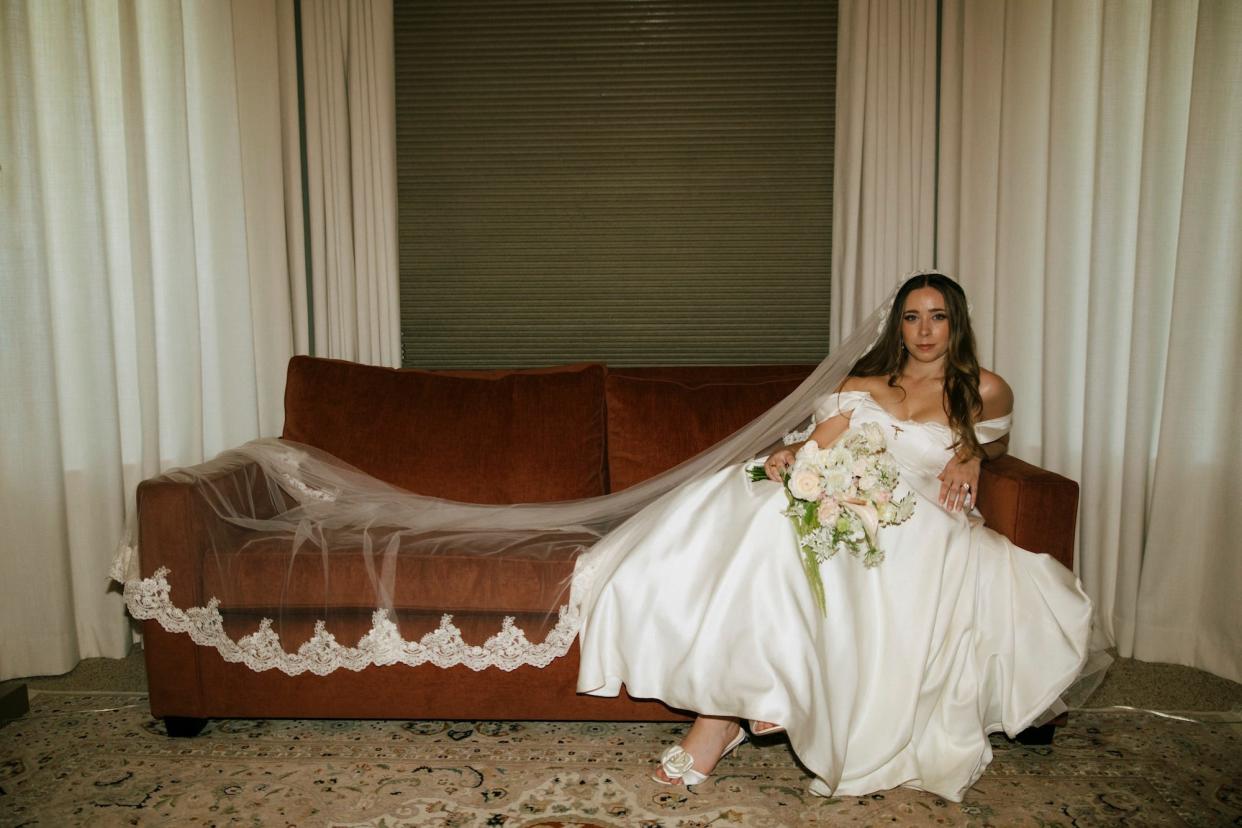 A bride sits on a couch with her veil flowing over it, wearing shoes with rosettes and holding a bouquet.