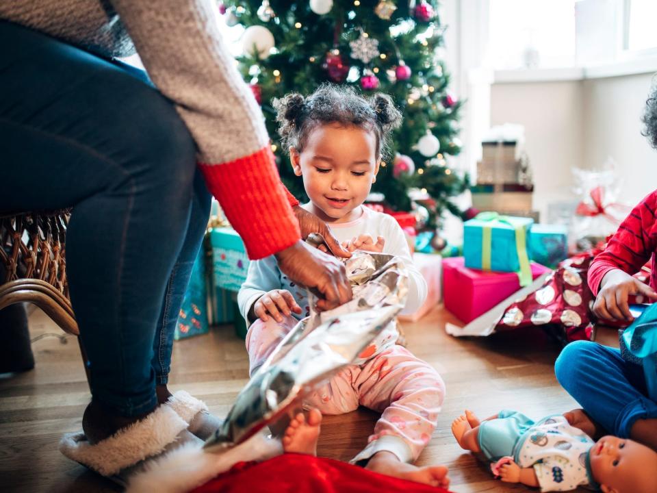 A parent helps a little girl open her Christmas presents as she sits on the floor.