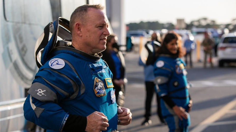 a man with short hair stands on the left, facing right, wearing a blue spacesuit, with the helmet unzipped around the collar and draped back like a hoodie. Another astronaut, out of focus, stands to his side, upward and to the right in the image. Par of a bus can be seen behind them, and crowds standing at the edge of a parking lot in the background.