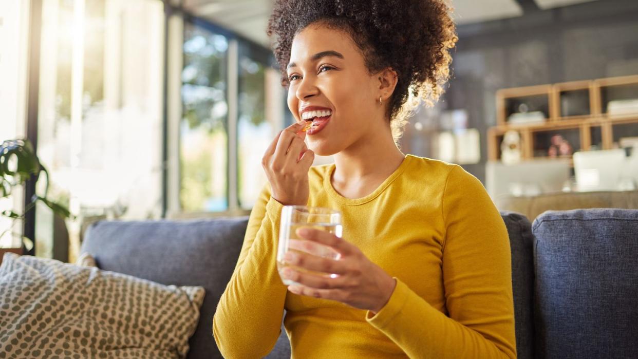 young happy mixed race woman taking medication with water at home one hispanic female with a curly afro taking a vitamin for good health while sitting on the couch at home woman drinking a supplement