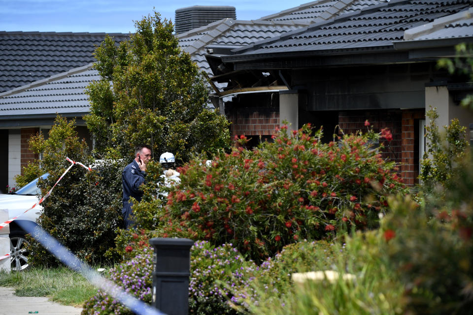 A red brick home with green bushes and flowers out the front and emergency services seen in the background.