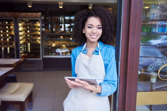 Young female coffee shop owner in apron at storefront, holding tablet.