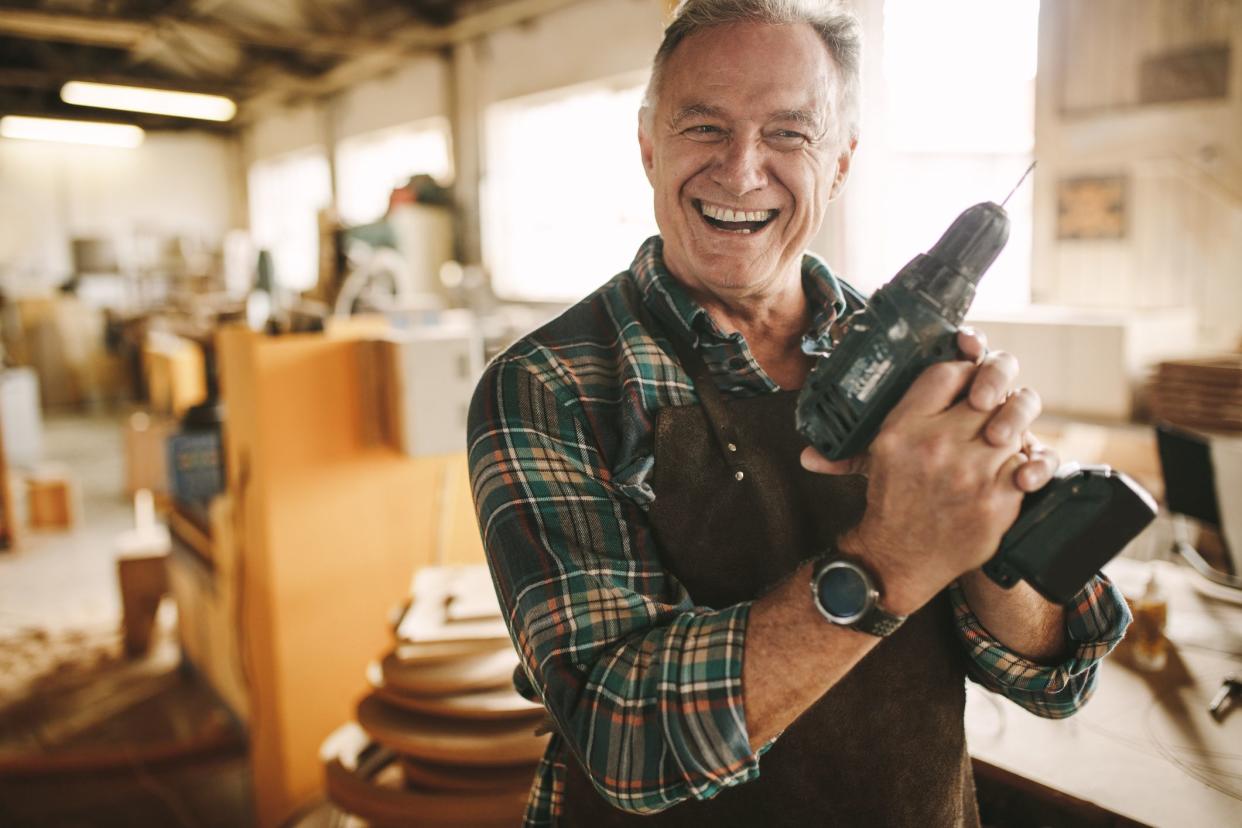 Smiling senior carpenter holding drill machine against workshop. Mature male worker smiling confidently to the camera holding drilling machine at his carpentry workshop.