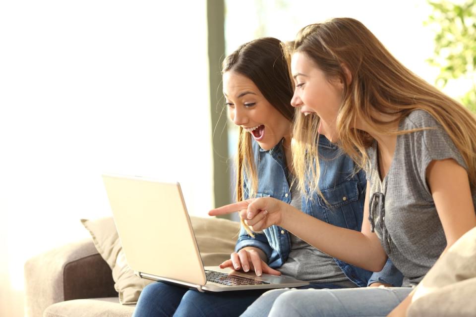 Two people smile while sitting on a couch and pointing at a laptop computer.