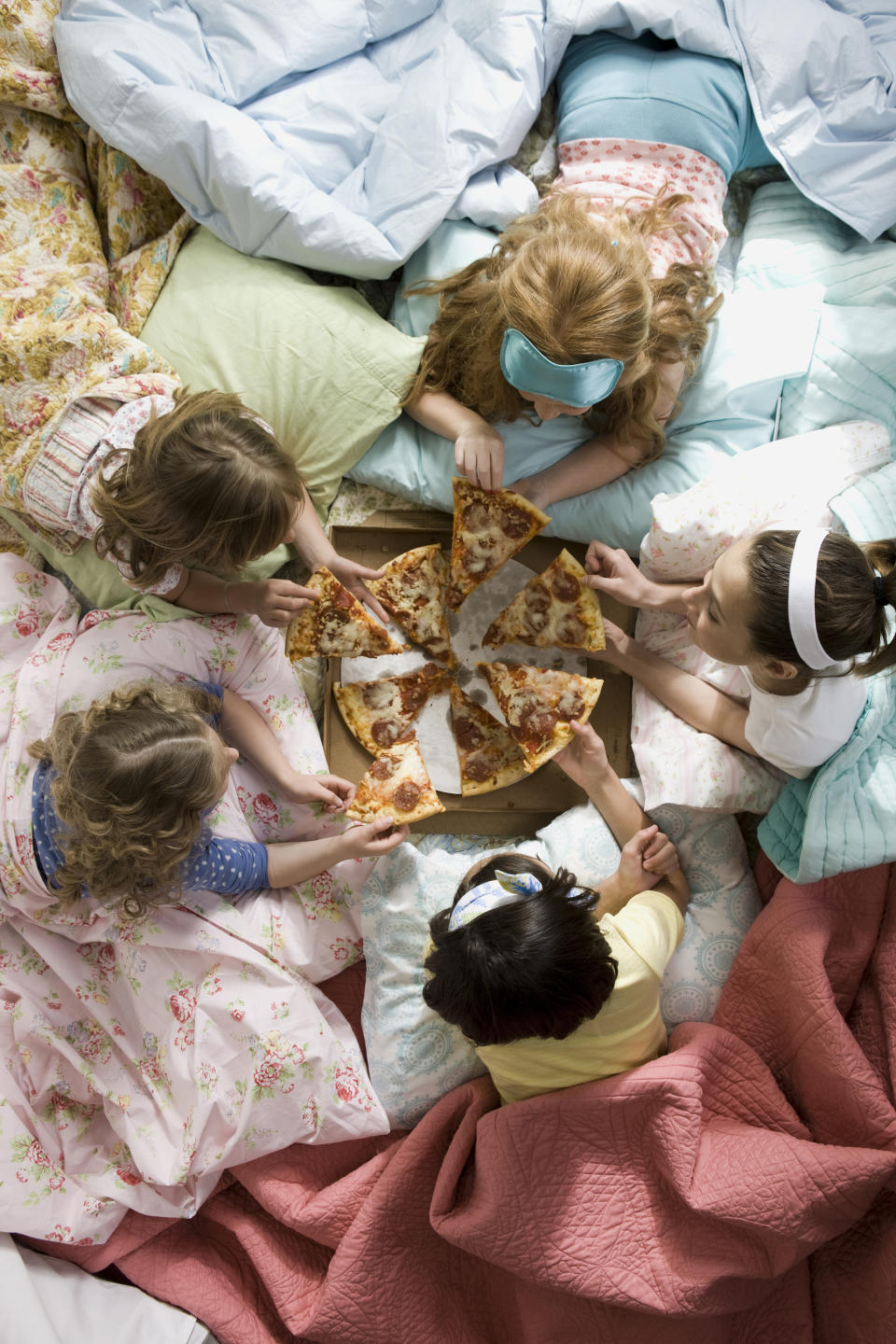 Five girls are cuddled up in blankets all facing each other, while they talk and eat slices of pepperoni pizza