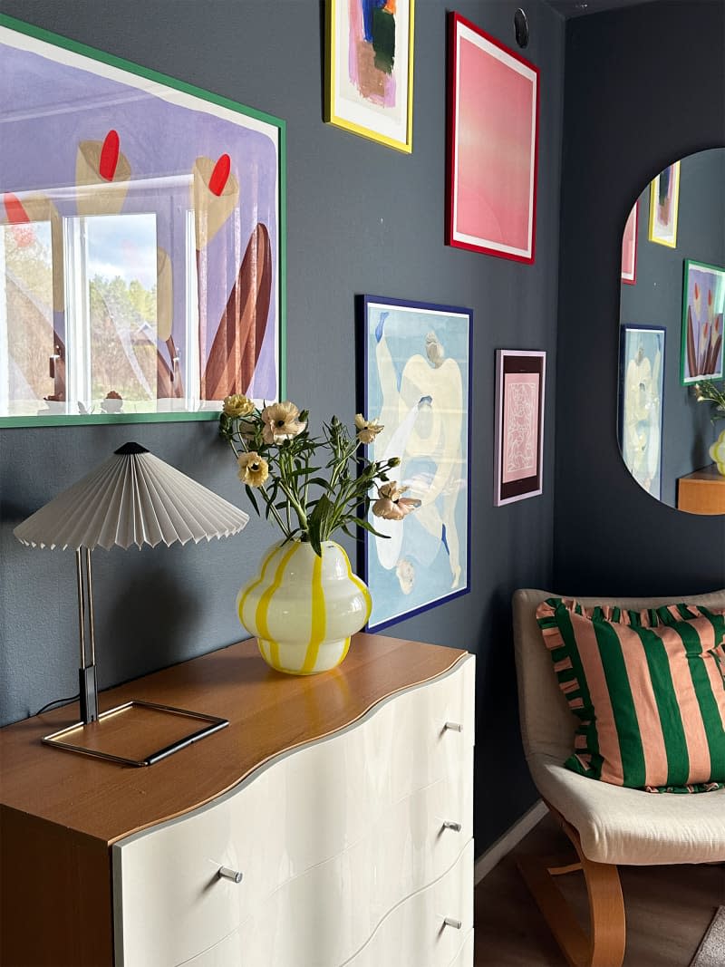 A multicolored vase sits on a dresser in a room filled with color and artwork.