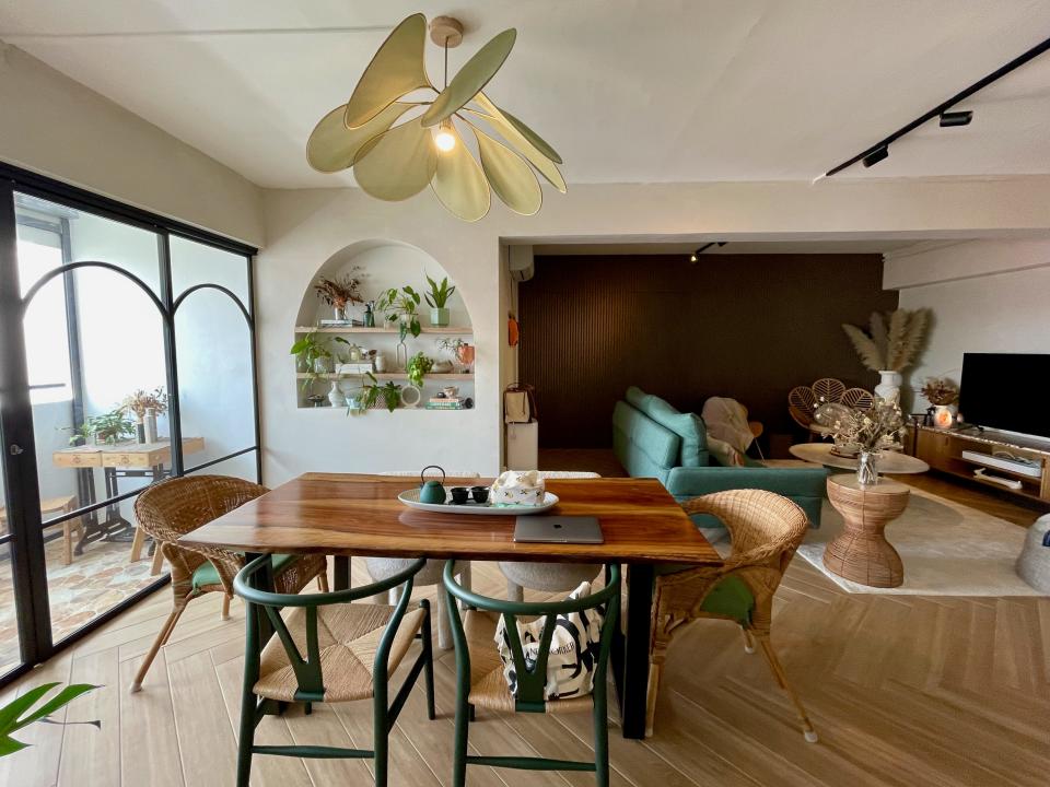 The open-plan living space in the couple's home, featuring a wooden dining table with wicker chairs, a shelf of plants, and a living space with couches and a TV off to the right.
