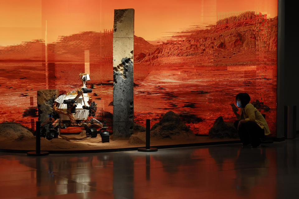 A woman wearing a mask talks on her phone near an exhibition depicting a rover in Mars in Beijing on Friday, May 14, 2021. China says its Mars probe and accompanying rover are to land on the red planet sometime between early Saturday morning and Wednesday Beijing time. (AP Photo/Ng Han Guan)