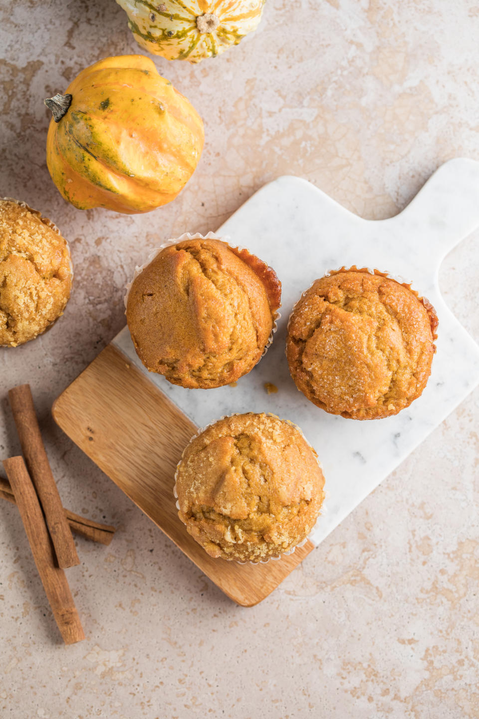 Four pumpkin muffins arranged on a marble and wood cutting board next to two pumpkins and cinnamon sticks