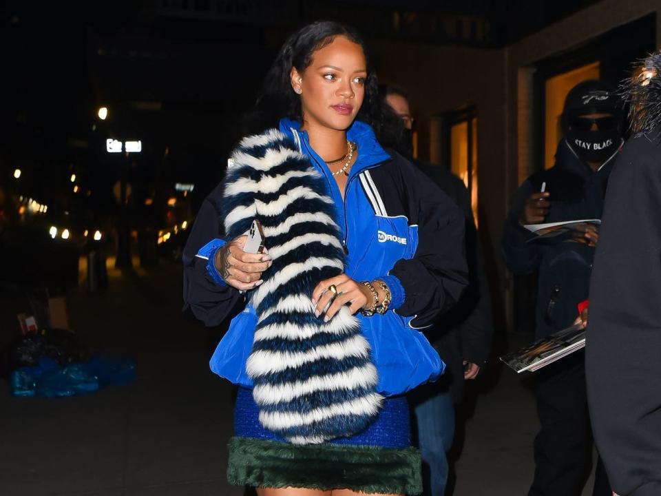 Rihanna in a blue and black windbreaker jacket with a black and white striped fur scarf over her shoulder. She's wearing a blue skirt with dark green trim and white heels with straps wrapped around her legs up to her knees.