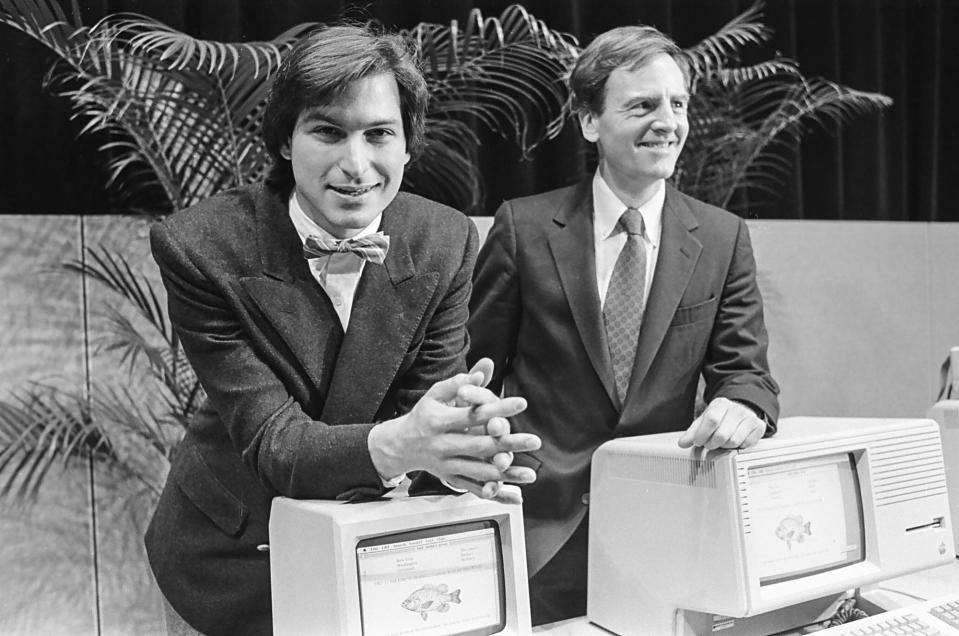 Steve Jobs and John Sculley lean on original Apple computers, Lisa and Macintosh, in a black and white photo