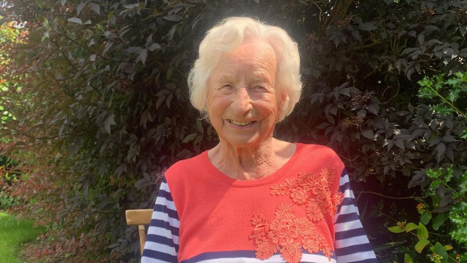 A smiling Joan Green looking directly at the camera and sitting on her chair in a garden, wearing a striped top. 