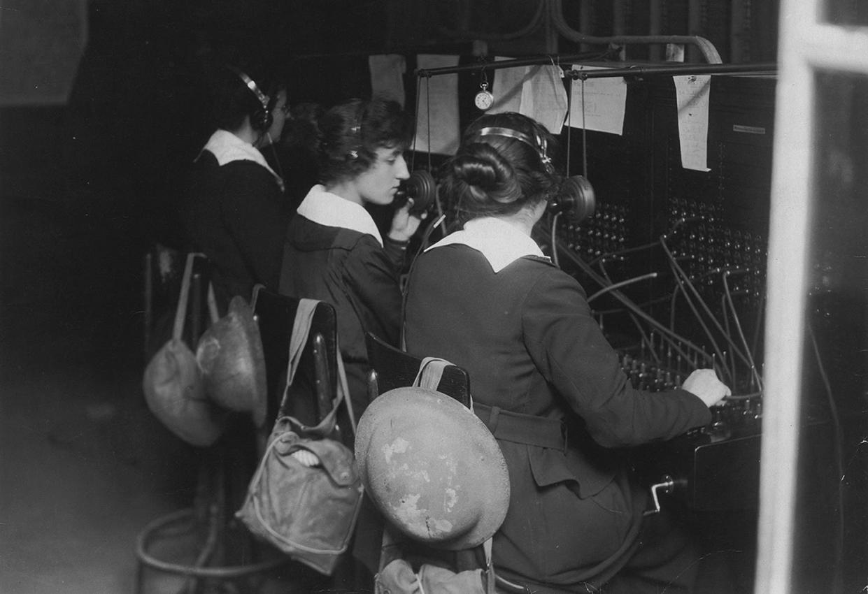 The Hello Girls work the switchboard on the frontlines of World War I. Strapped to the back of their chairs are trench helmets and gas masks.