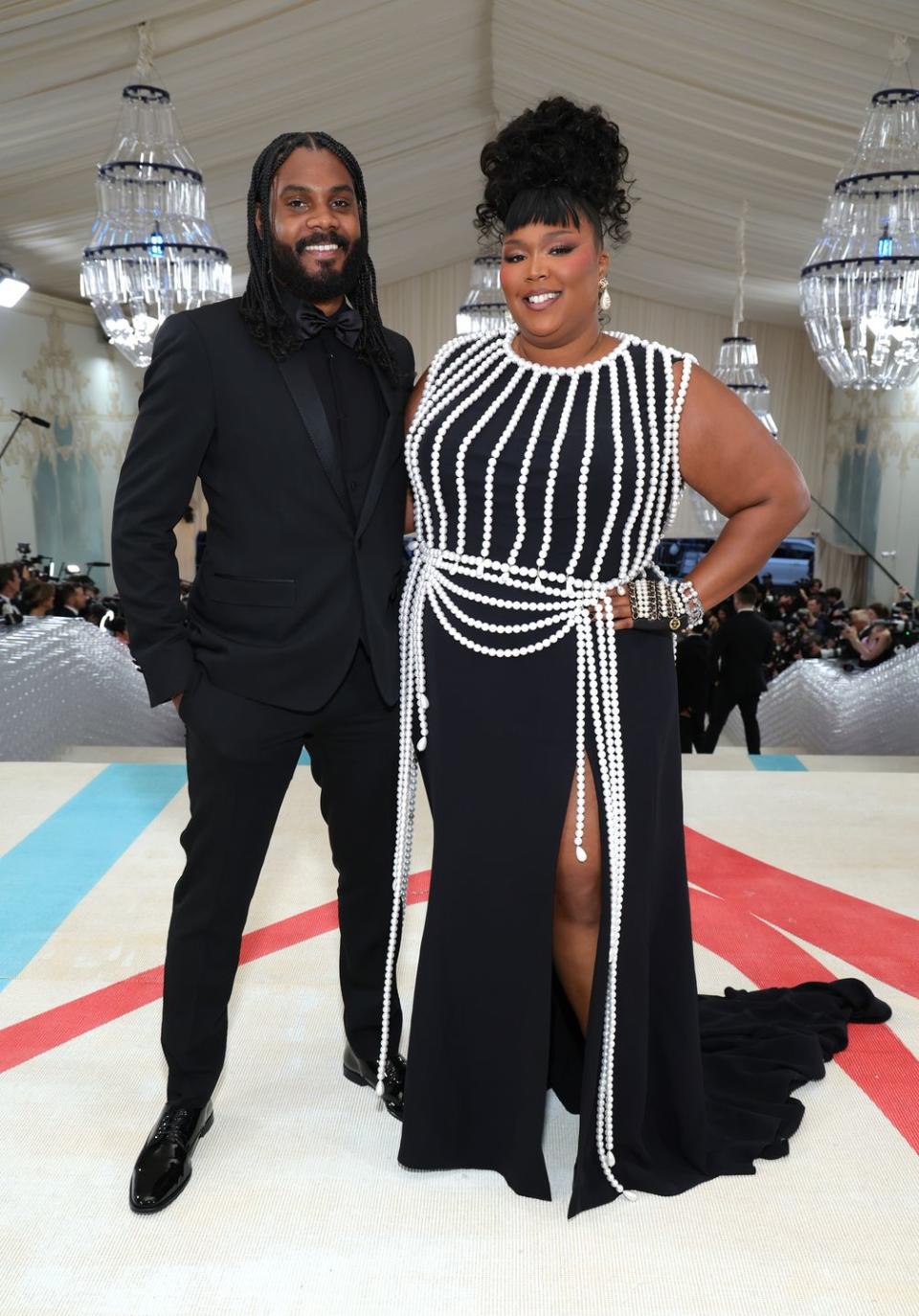 myke wright and lizzo stand next to each other embracing and smiling at the camera, he wears an all black tuxedo, she wears a black dress adorned with white pearls and pearl jewelry