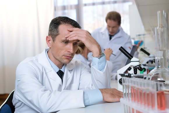 A disappointed-looking man in a lab coat sitting at a table with lab equipment.