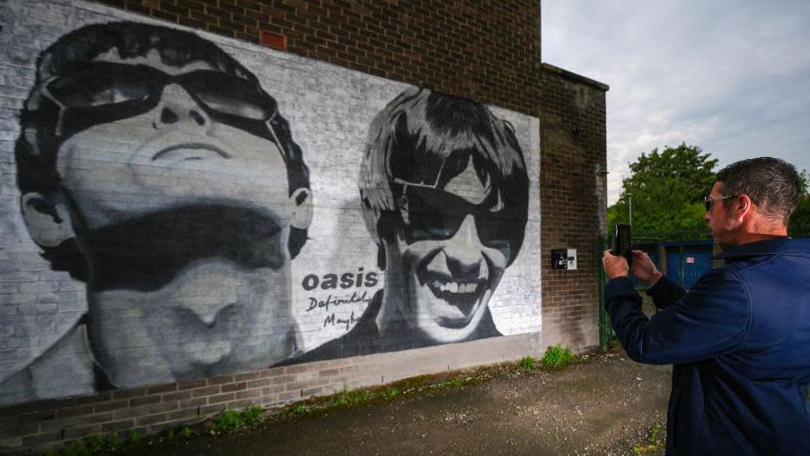 Oasis fan photographing a new mural of Liam and Noel Gallagher of Oasis, created by Manchester street artist Pic.One.Art., on the side of the Sifters Record store in Burnage, in Manchester